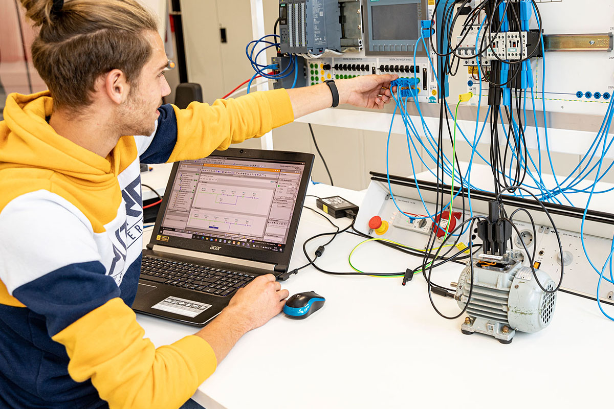 Auszubildender arbeitet mit Notebook und verdrahtetem SPS-Schulungsaufbau an einer Automatisierungsanlage.