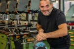 Person arbeitet an einem Schraubstock in einer Metallwerkstatt mit mehreren Standbohrmaschinen im Hintergrund.