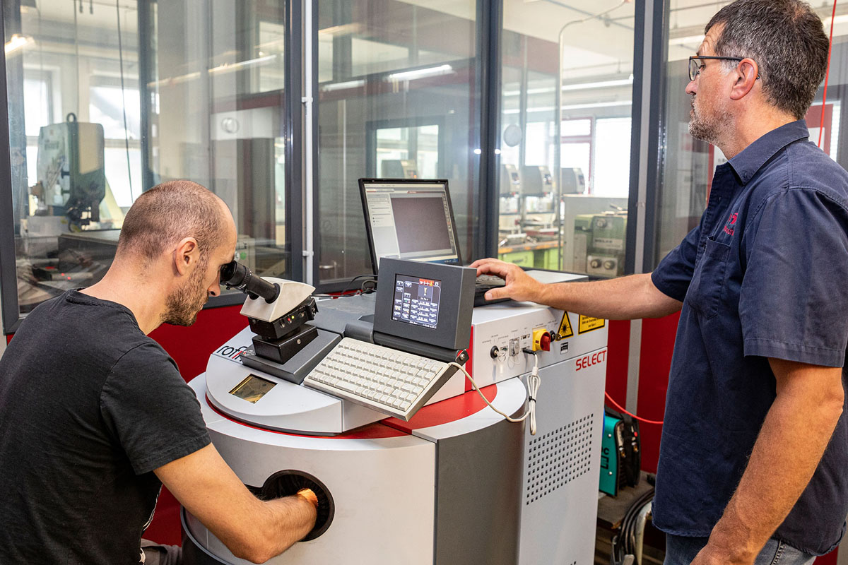 Teilnehmer arbeitet an einer Laserschweißanlage mit Mikroskop, während eine weitere Person Einstellungen am System vornimmt.