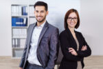 Zwei Personen in Business-Kleidung stehen vor einem Flipchart in einem Seminarraum.