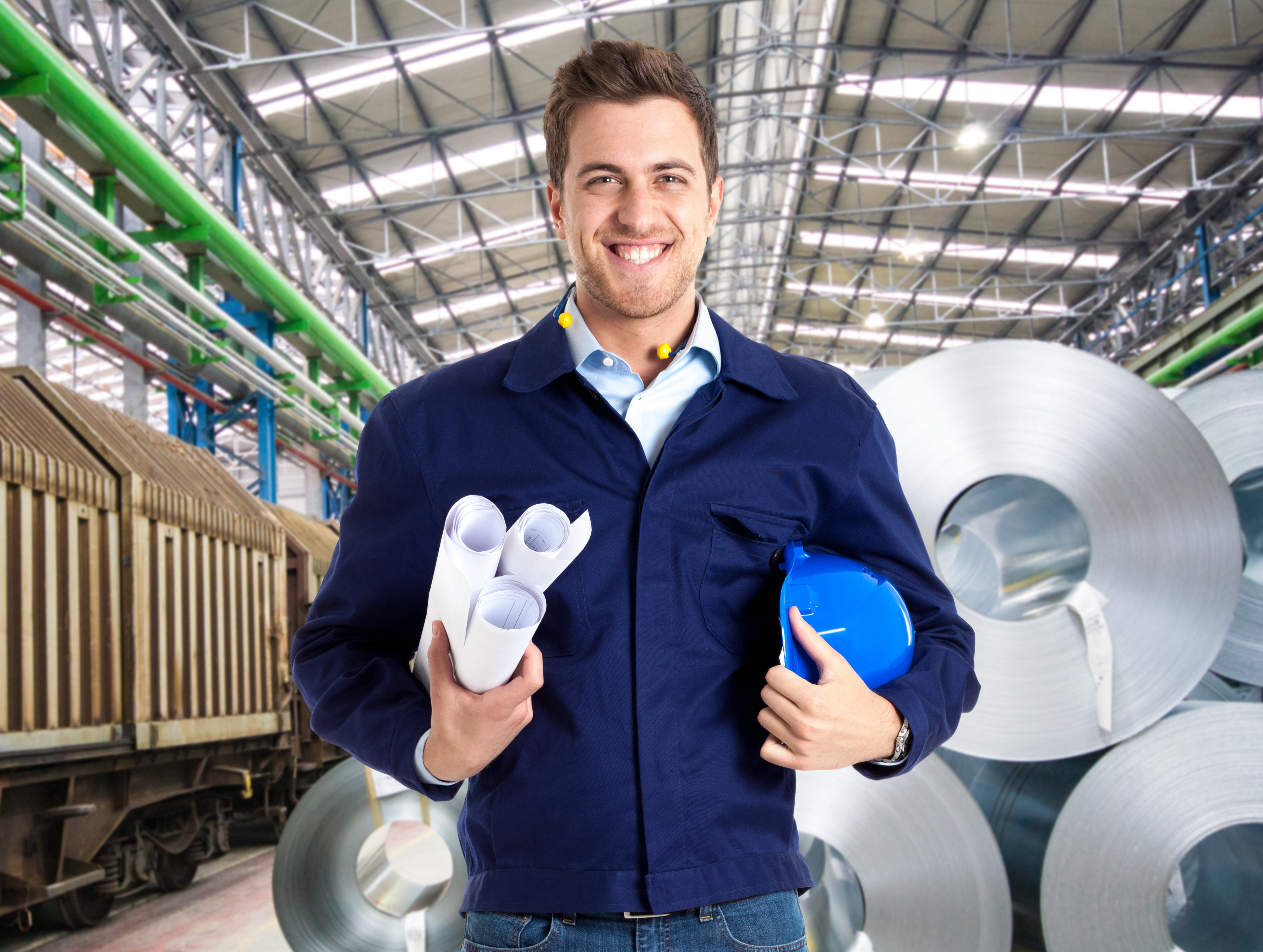 Person in Arbeitskleidung hält Baupläne und Schutzhelm in einer Produktionshalle mit Metallcoils.