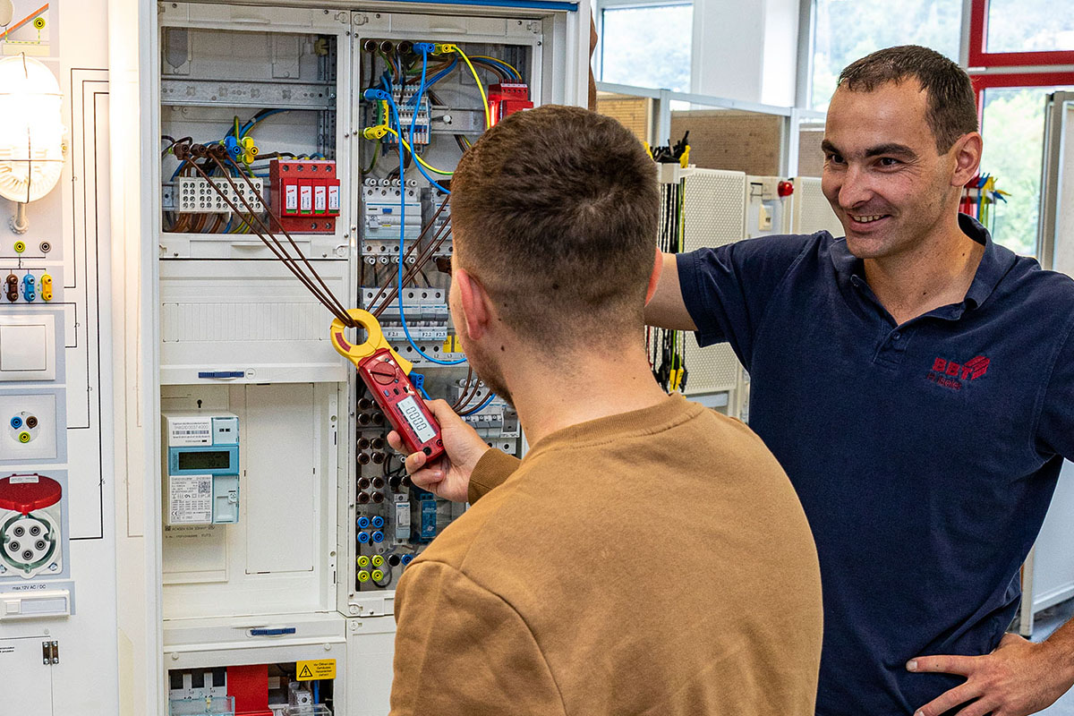 Teilnehmender misst an einer geöffneten elektrischen Schaltanlage, während eine weitere Person die Arbeit begleitet.