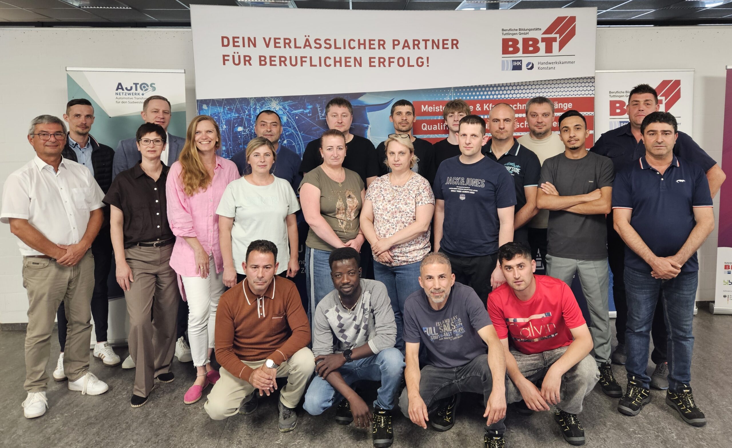16 Absolventinnen und Absolventen der Qualifizierungsmaßnahme Fachkraft in der Produktion mit Vertretern von BBT und Vatter Bildungszentrum vor BBT-Banner