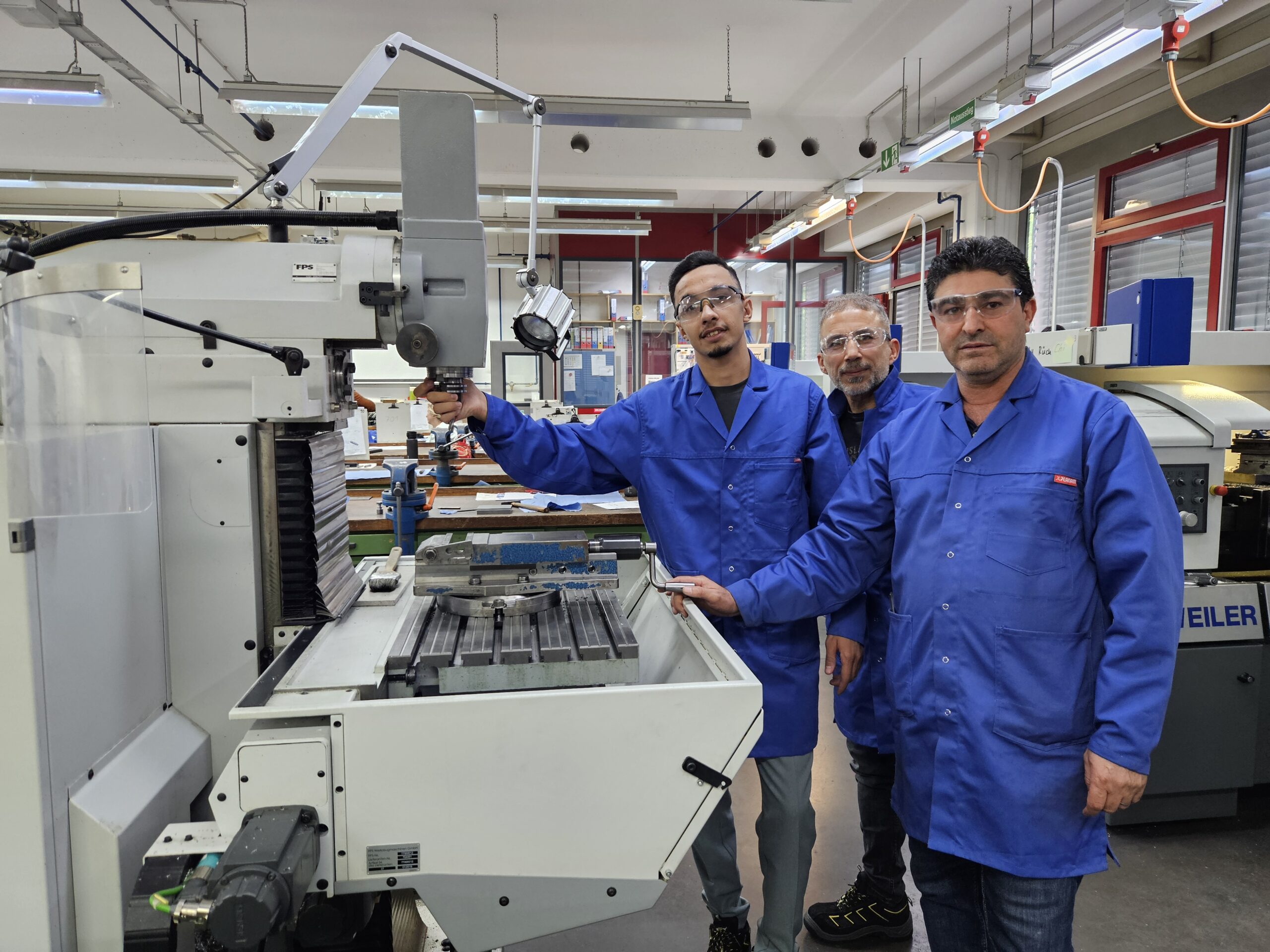 Drei Teilnehmende der Qualifizierungsmaßnahme Fachkraft in der Produktion in blauen Arbeitskitteln an einer Werkzeugmaschine in der BBT